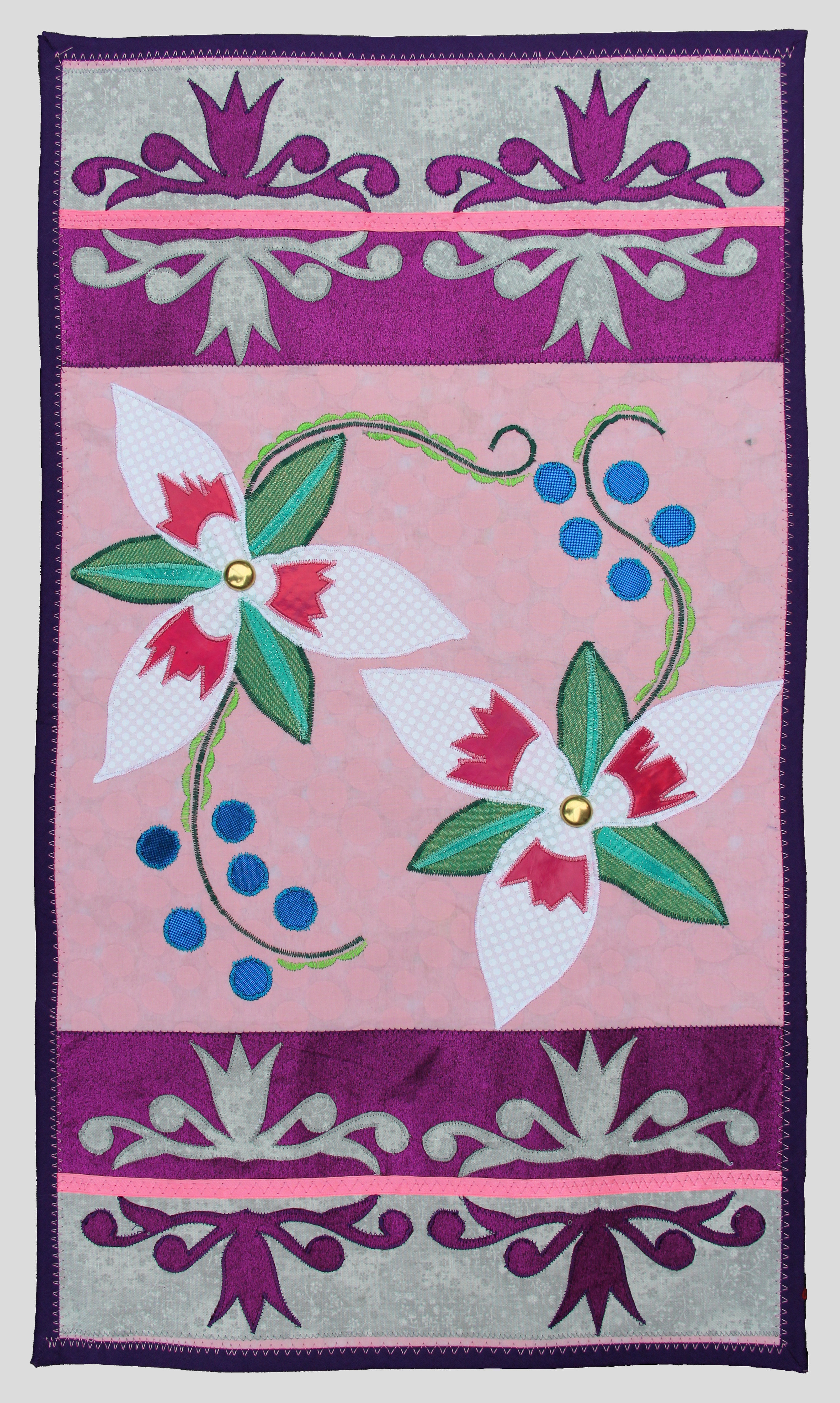 A rectangular textile hand made by Angela Waupochick for the Nisinoon project, displaying a top-down view of two trillium flowers at opposite corners of a pink square in the center, bordered by side views of two trillium flowers along the top and bottom. Photo by Rodney Schreiner.